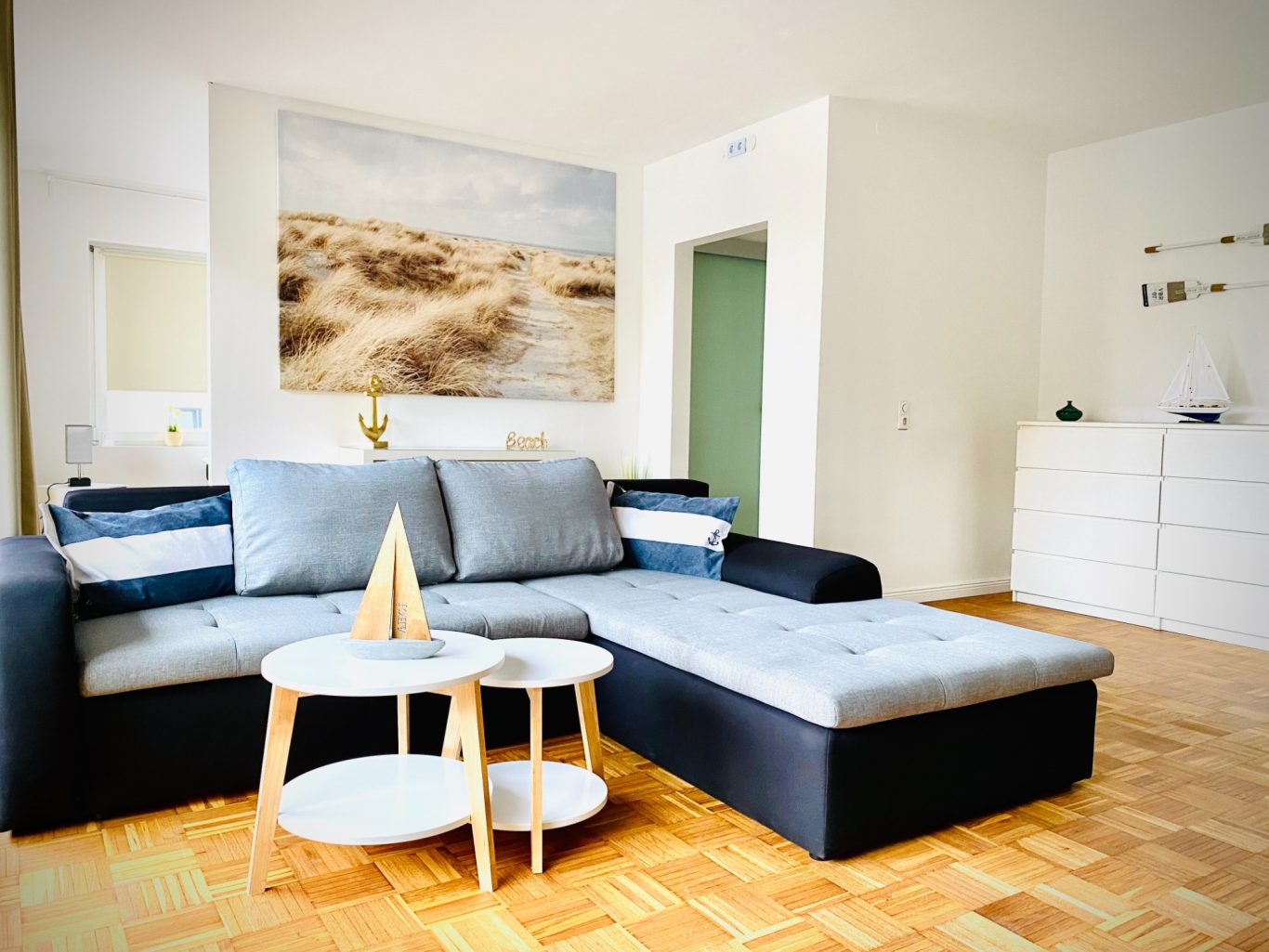Heller Wohnbereich mit Schlafsofa in der Wohnung Meeresbrise Helles Wohnzimmer mit grauem Sofa, Couchtischen und Landschaftsbild an der Wand in Sierksdorf